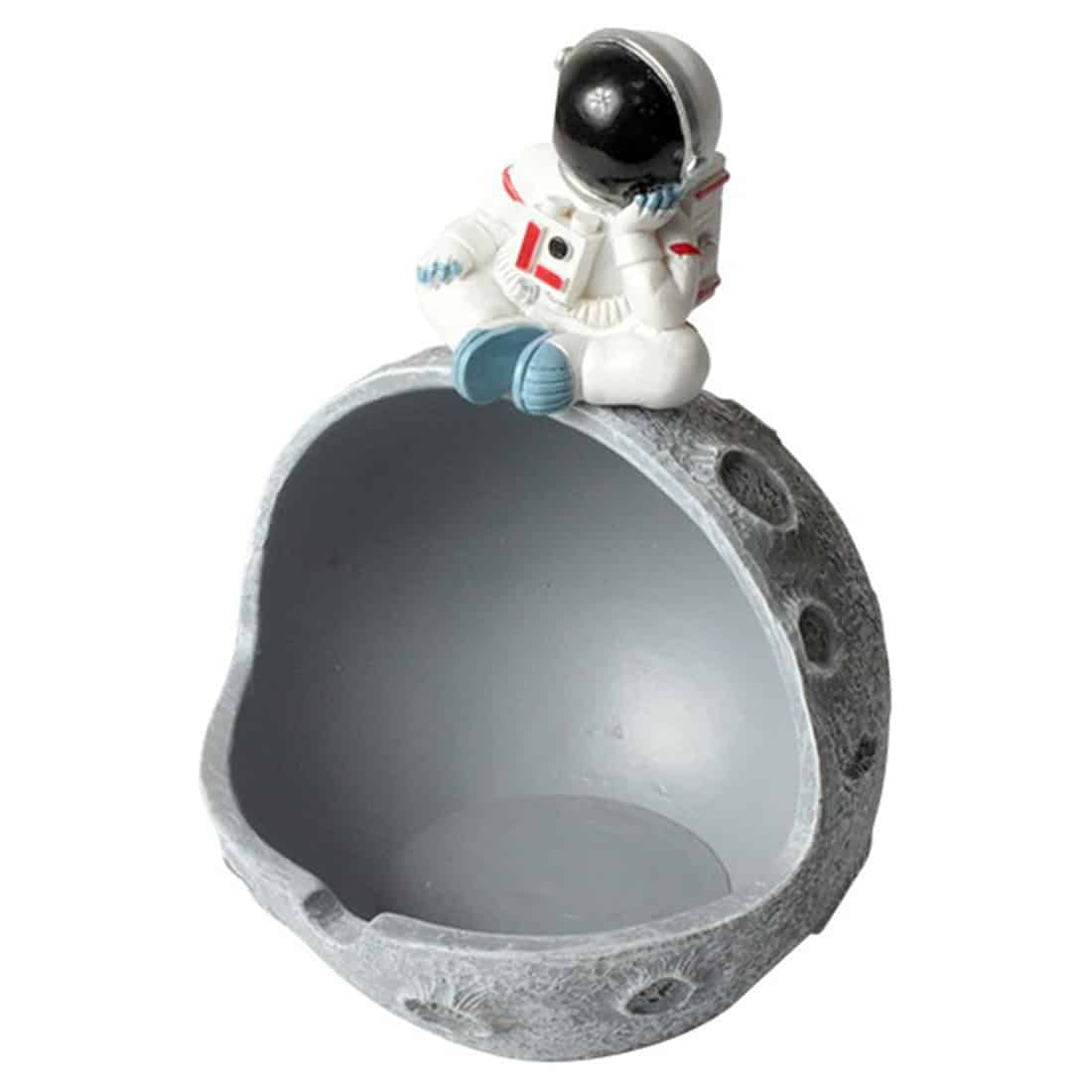 En ashtonaut-ashtray præsenterer en detaljeret figur af en astronaut i en lys grå farve, iført en rumdragt med synlige teksturer og sølvfarvede detaljer. Astronauten sidder med benene let adskilt, og en halvmåneformet skål er placeret på det nederste segment, hvor der er plads til aske. Baggrunden er en simpel, ensfarvet overflade, der fremhæver figuren.