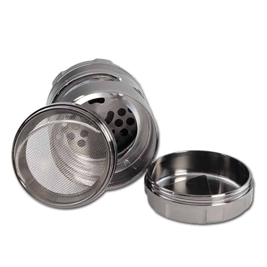 En metalgrinder med en grå overflade og en diameter på 63 mm. Grinderens krop har en børstet metalfinish, der giver en mat tekstur. Der er synlige skarpe tænder indeni, der er designet til at male krydderier eller urter. Låget har et let skrue- eller kliksystem, som sikrer en tæt lukning.