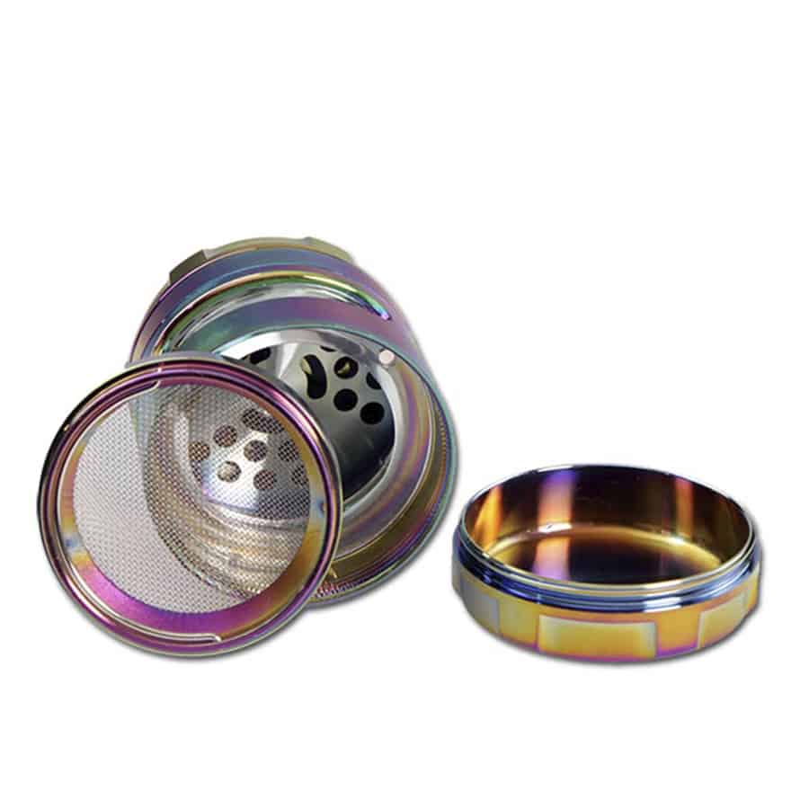 En metalgrinder med en olie-farvet overflade præget af dybe, glatte nuancer af grøn og blå. Den har en cylindrisk form med en diameter på 63 mm. Overfladen er blank og reflekterer lys, hvilket skaber et skinnende udtryk. Grinderens top har en skridsikker tekstur med små, ergonomiske knopper for et bedre greb. Indeni ses skarpe tænder til formaling.