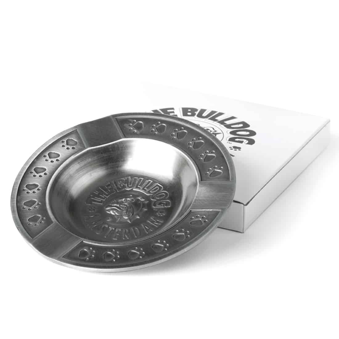 En firkantet askebæger i metal med en mat, grå overflade. Askebægeret har en præget tekstur med et bulldog-motiv i midten, der tilføjer dybde. Randen er let hævet, hvilket skaber en tydelig afgrænsning. Der er fire små indhak til cigaretter langs kanten, hvilket giver funktionel plads. Materialet fremstår holdbart og robust.
