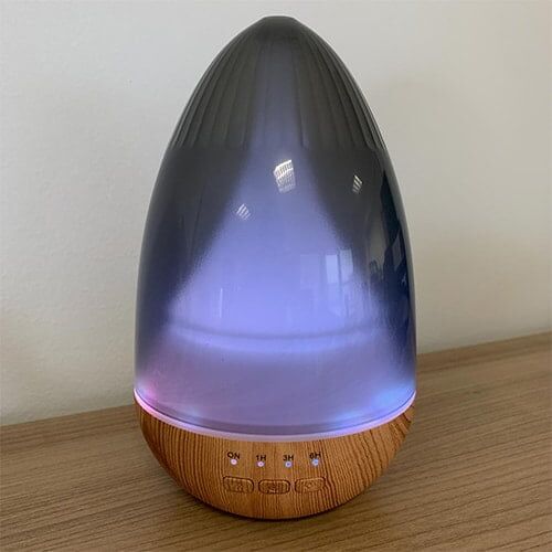 En cylindrisk aroma diffuser i naturligt trælook med en glat overflade. Den øverste del er buet og har en let skråning mod midten, hvor der er en åbning til dampudslip. Diffuseren har en USB-port i bunden og er omgivet af en lys ring, der kan skifte farve. Mønstret på træoverfladen er subtilt og varieret.