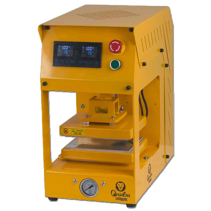 En robust, metallisk presse med en dybgrå overflade pryder midten af scenen. Den har en stor, firkantet varmeplade øverst, der er glat og blank. På forsiden findes kontrolknapper i sort med tydelige symboler. Pressebenene er solide og giver stabilitet. Bagpå er der tilslutningsporte, der understøtter funktionaliteten. Hele strukturen udstråler holdbarhed og styrke.