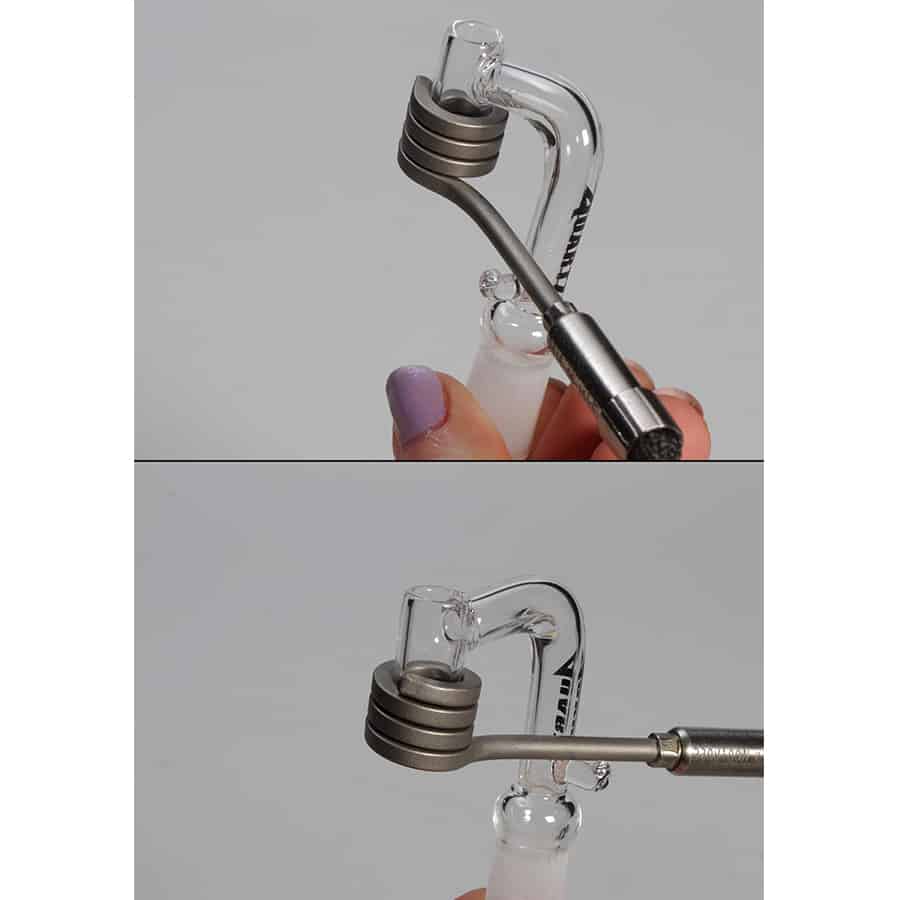 En kvarts dab banger i gennemsigtig, klart materiale pryder midten af kompositionen. Den har en cylindrisk form med en bred åbning for oven og en smalere bund. En varmelegeme er synligt monteret på siden, og der er en lille pod, der tilsluttes. Overfladen er glat med lette refleksioner, og farven er næsten farveløs med en let skygge.