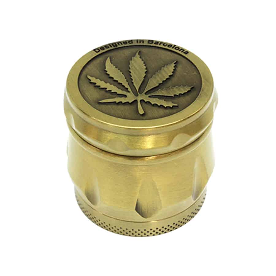 En guldmalet krydderikværn pryder overfladen med et præg af blade og detaljeret mønster, der giver en taktil kvalitet. Overfladen reflekterer lys med en glinsende finish. Kværnen har en rund form med en justerbar top og en glat, ensartet tekstur, der står i kontrast til de dybe detaljer i prægningen. Den er placeret på en neutral baggrund, hvilket fremhæver dens design.