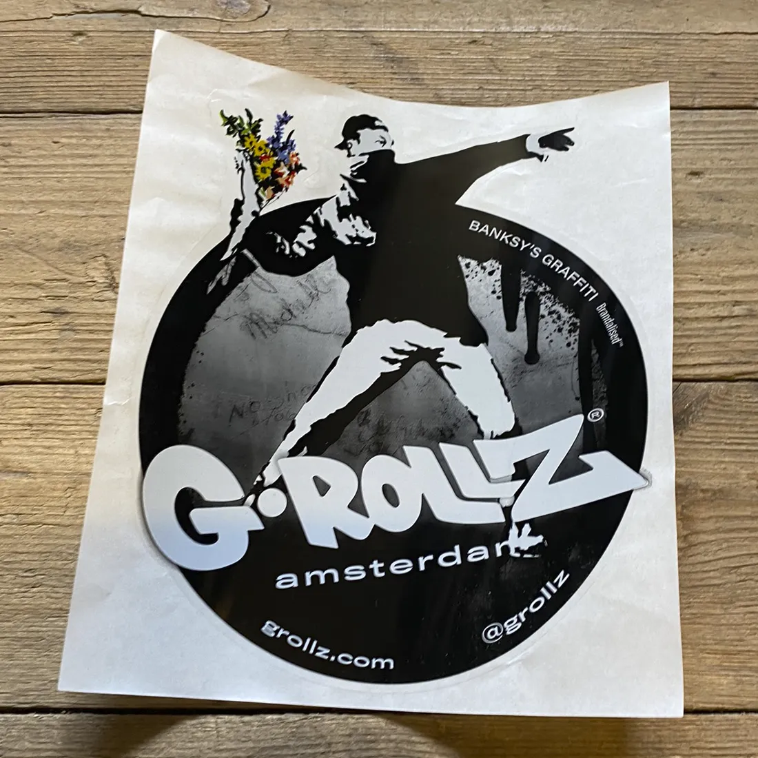 Klistermærket præsenterer et grafisk design med en blomsterbuket, hvor blomsterne er i nuancer af lilla, gul og hvid. Blomsterne er arrangeret i en vilkårlig komposition, der skaber bevægelse. Baggrunden er i en lys tone, der fremhæver farverne. Der er også et subtilt mønster, der tilføjer tekstur til det samlede udtryk.