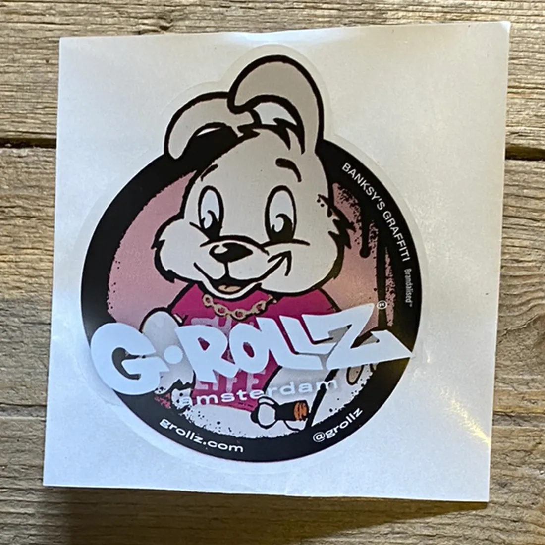 Klistermærket præsenterer en grafisk illustration af en kanin i sort og hvid, iført en hætte og med en pistol i hånden. Baggrunden er en blanding af grå og hvid nuancer, der giver en rå effekt. Kaninens ansigtstræk er detaljerede med klare konturer, mens teksten "G-Rollz" er placeret nederst med en moderne skrifttype.