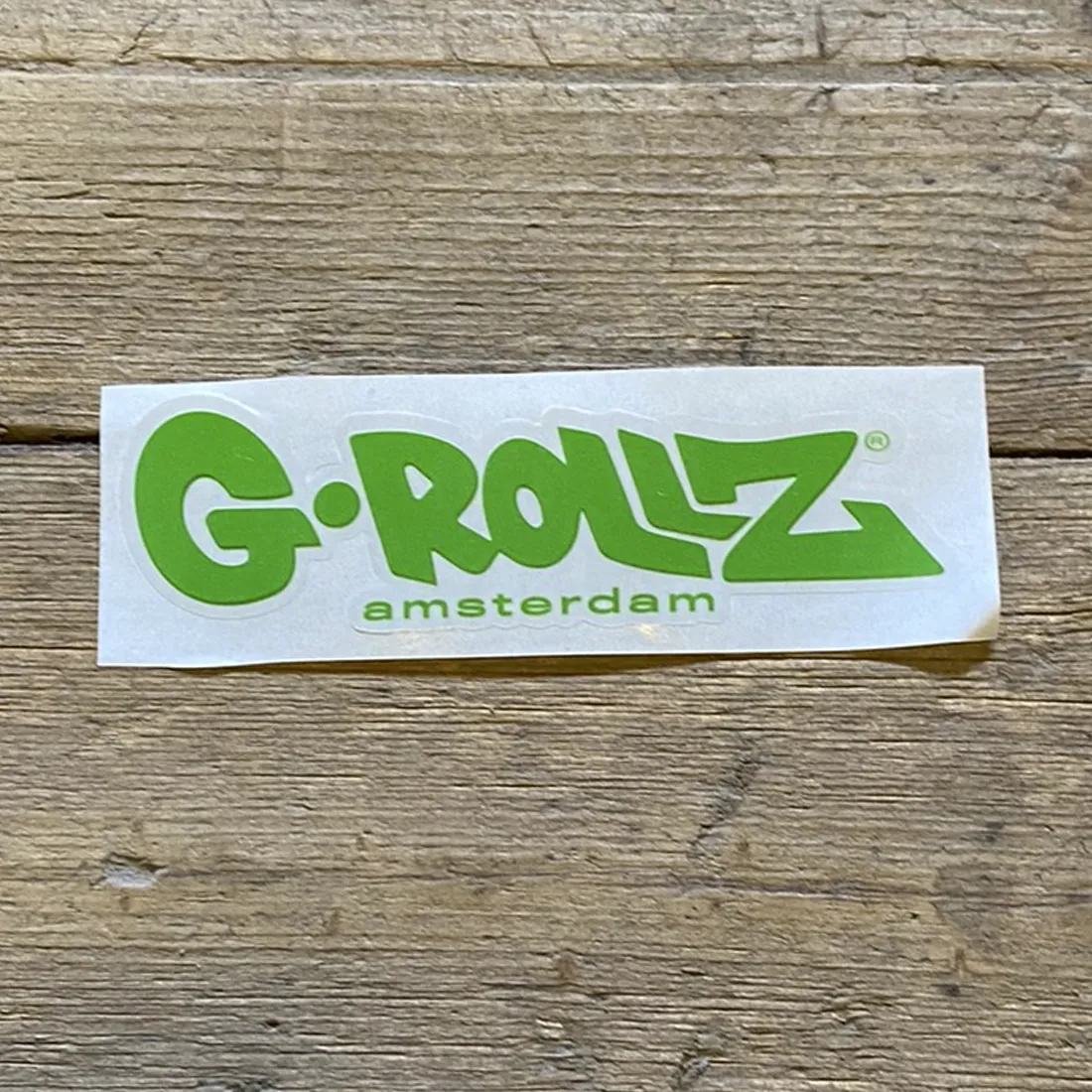 Klistermærket har en rund form med en klar, grøn baggrund. I midten fremtræder et hvidt logo, der består af en stiliseret tekst med en moderne skrifttype. Det grønne område er jævnt og glansfuldt, hvilket giver en skarp kontrast til den hvide tekst. Omkring kanten er der en subtil, hvid kantlinje, der fremhæver designet.