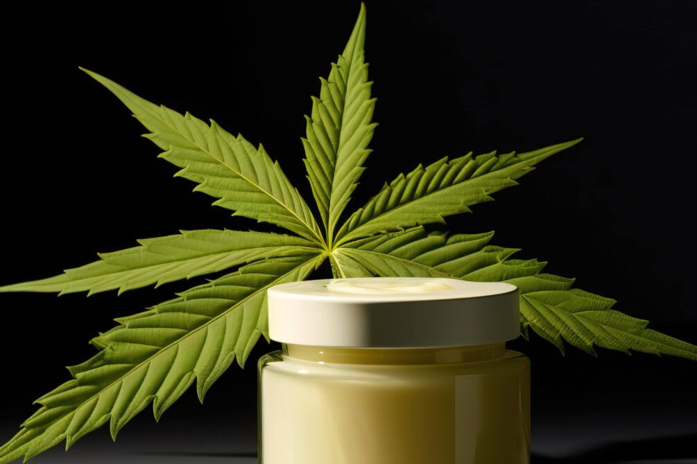 En rund, klart glas krukke med et sort låg indeholder en grønlig, cremet substans. Krukken står på en lys træoverflade, hvilket fremhæver dens glans. Substansen har en ujævn tekstur med små stykker, der er synlige. Baggrunden er neutral og uklar, hvilket skaber fokus på krukken og indholdet.