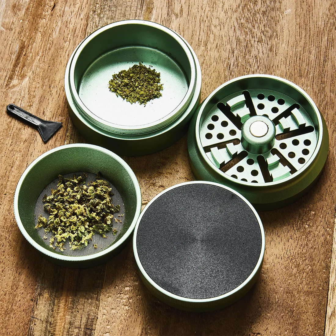 En grøn, rund krydderkværn med en diameter på 55-63 mm. Overfladen har en mat finish, mens kanten har en glat, poleret tekstur. Kværnen er opdelt i flere sektioner, hvor den øverste del har små tænder til formaling. Der er en synlig, skridsikker ring langs bunden, og en lille åbning til udskillelse af det formede indhold.