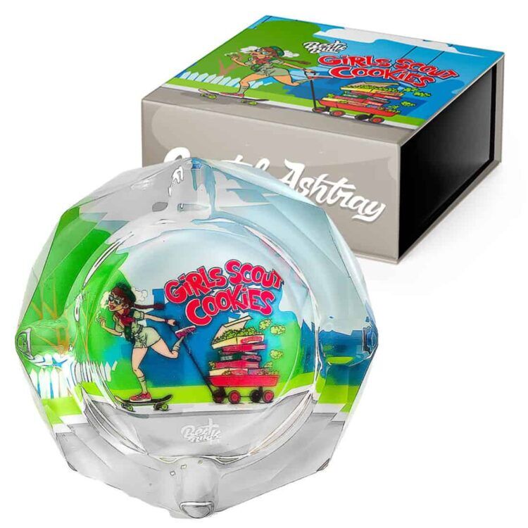 En rund, klar krystalaskebæger med en glat overflade og skarpe kanter prydes af en indgraveret tekstur, der skildrer to stiliserede cannabisblade. Askebægeret har en let vægt og en æstetisk, minimalistisk design, der fremhæver klarheden i materialet. Refleksioner fra lys skaber en glitrende effekt, der tilføjer dybde til objektet.