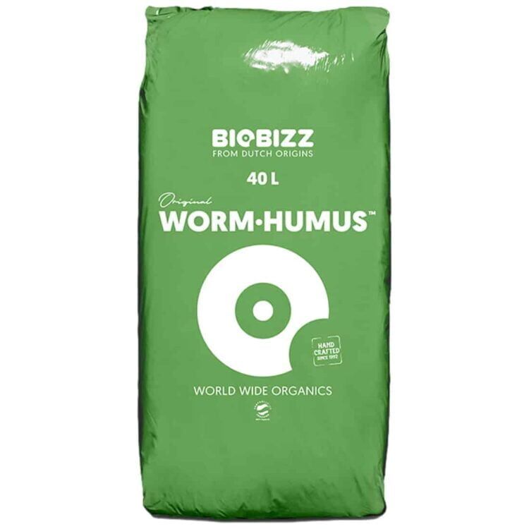 Mørkebrunt, granuleret materiale fylder en stor, transparent plastikpose. Overfladen er ujævn med små klumper, der skaber en grov tekstur. Posen er lukket med en sort zip-lukning, og der er et farvetiket på forsiden med grønne og blå nuancer. Baggrunden er neutral, hvilket fremhæver posens indhold.