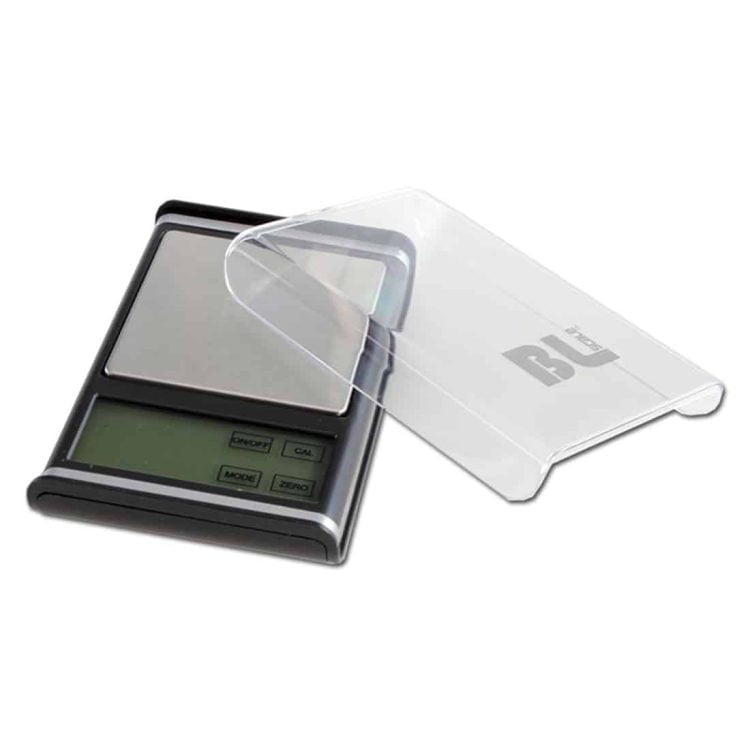 En skala-model af et hus præsenteres med en præcis detaljeret struktur. Taget har en skrå form med mørkt tagpap, mens væggene er i lys gråfarve med vinduer i hvid ramme. Terrænet omkring modellen er dækket af grønne græsstrå, og der er små træer i forskellige nuancer af grøn. Modellen står på en neutral, hvid overflade.