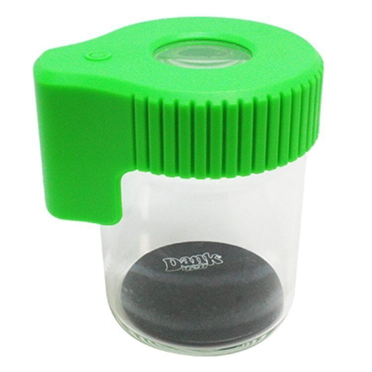 Forstørrelsesglasset har en rund, grøn krukke med en glat overflade og en let gennemsigtig struktur. Låget er også grønt og har en flad, cirkulær form med en skarp kant. Forstørrelsesglasset, der er placeret oven på krukken, har en klar linse og en kraftig, hvid plastikramme, der giver en solid kontrast til den grønne farve.