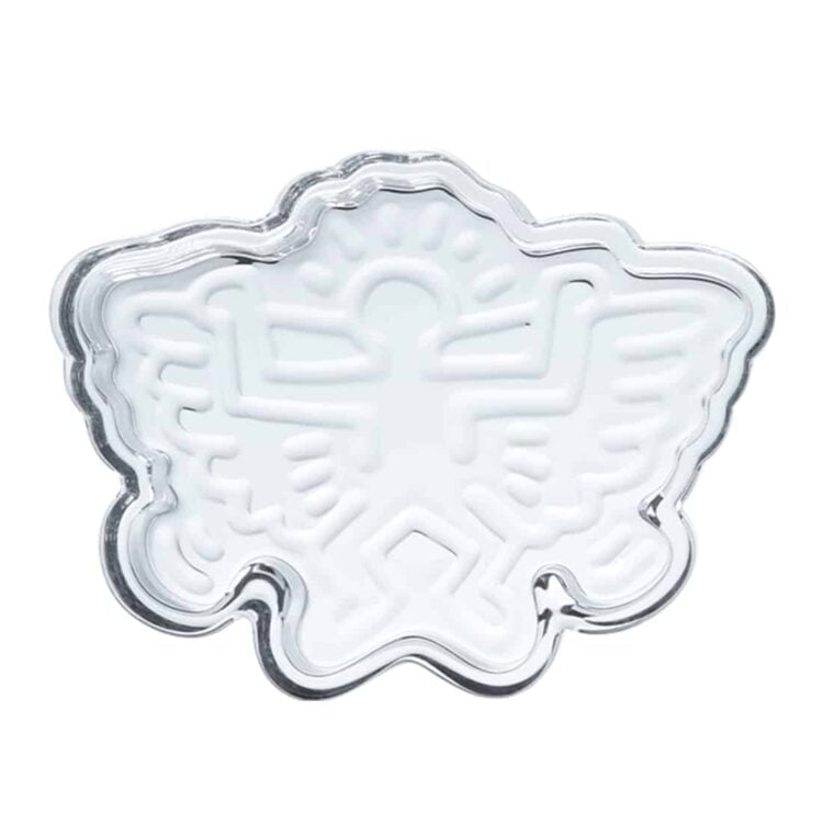 En flad skål i en lys, grålige nuance med en glat overflade pryder midten. Skålen har en let opadgående kant og en uregelmæssig form, der giver et organisk udtryk. Indvendigt ses et subtilt mønster, der tilføjer tekstur. Et lille, elegant håndtag i form af en engel er placeret på kanten, fremhævet med fine detaljer.