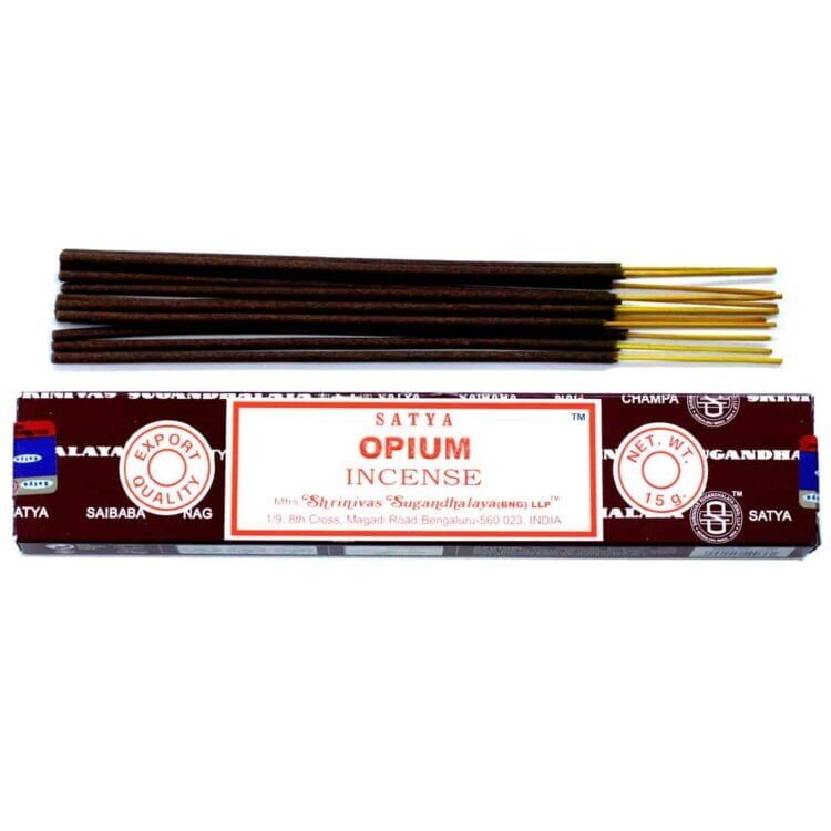 En pakke med røgelse præsenteres i en rektangulær, farverig indpakning. Forsiden prydes af et mønster med blomster og spiraler i nuancer af lilla, gul og grøn. Teksten er trykt i hvid og sort, med tydelige symboler. Røgelsen er pakket i en gennemsigtig plastpose, der viser de små stænger i naturlige toner af brun.