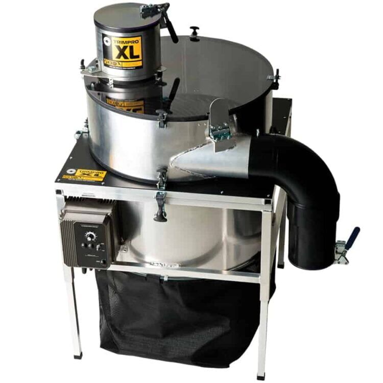 En stor, firkantet maskine med en robust metalramme dominerer kompositionen. Den har en glat, sort overflade med nogle sølvfarvede detaljer og betjeningsknapper. Fronten rummer en gennemsigtig beholder, hvor en fyldig, grøn plante ses. Maskinen står på en flad, hård overflade, og der er en elektrisk ledning, der strækker sig fra den.