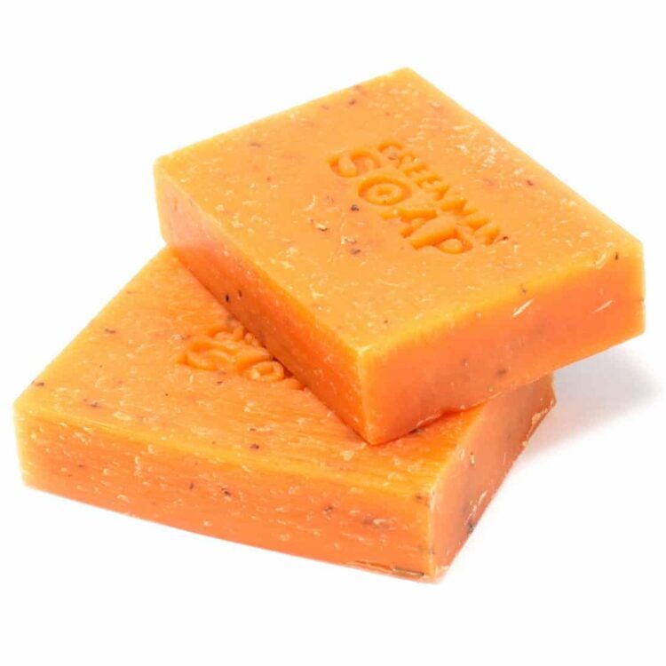 En firkantet sæbe med en glat, lys grøn overflade pryder midten af kompositionen. Sæben vejer 100 gram og er indpakket i en transparent plastfolie, der giver et glimt af den glinsende tekstur. På forsiden står der i elegant, gylden skrift "Golden Argan". Baggrunden er neutral, hvilket fremhæver sæbens farve og design.