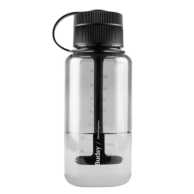 En gennemsigtig, grøn flaske med en afrundet bund og en smal hals pryder overfladen. Flaskens struktur har en let bølget tekstur, der giver et unikt greb. En indbygget skub-låg mekanisme sidder øverst, og en lille, buet mundstykke er placeret på siden. Farven skifter fra klar til dyb grøn mod bunden, hvilket skaber en visuel dybde.