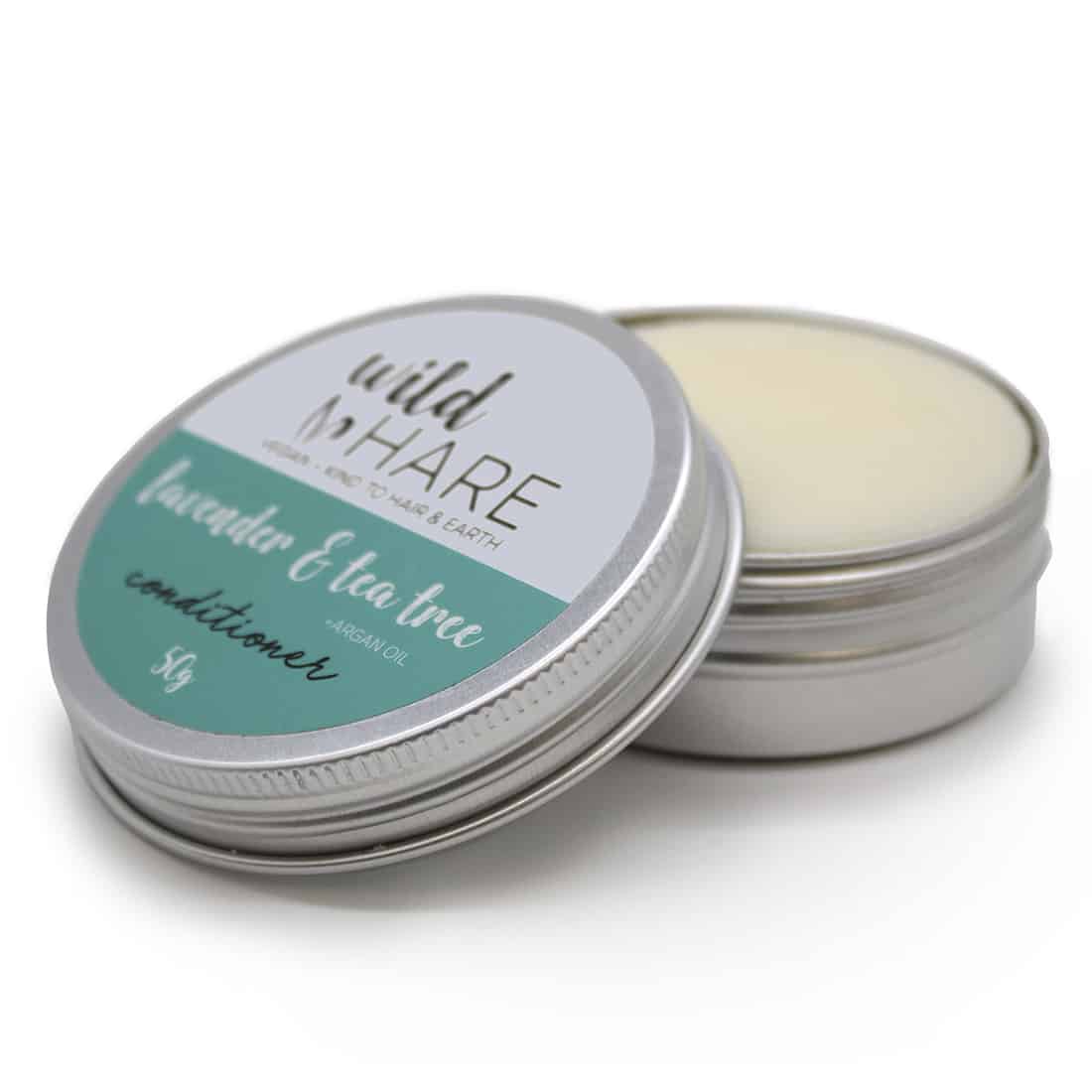 En rund, solid conditioner i en lys lavendelnuance, præget med en glat overflade. Den er indpakket i en papæske med et minimalistisk design, der fremhæver produktets naturlige ingredienser. Æsken har en mat finish med grønne og mørkegrønne skrifttyper, der angiver indholdet, herunder tea tree og lavendel. Baggrunden er neutral, hvilket fremhæver produktets farver.