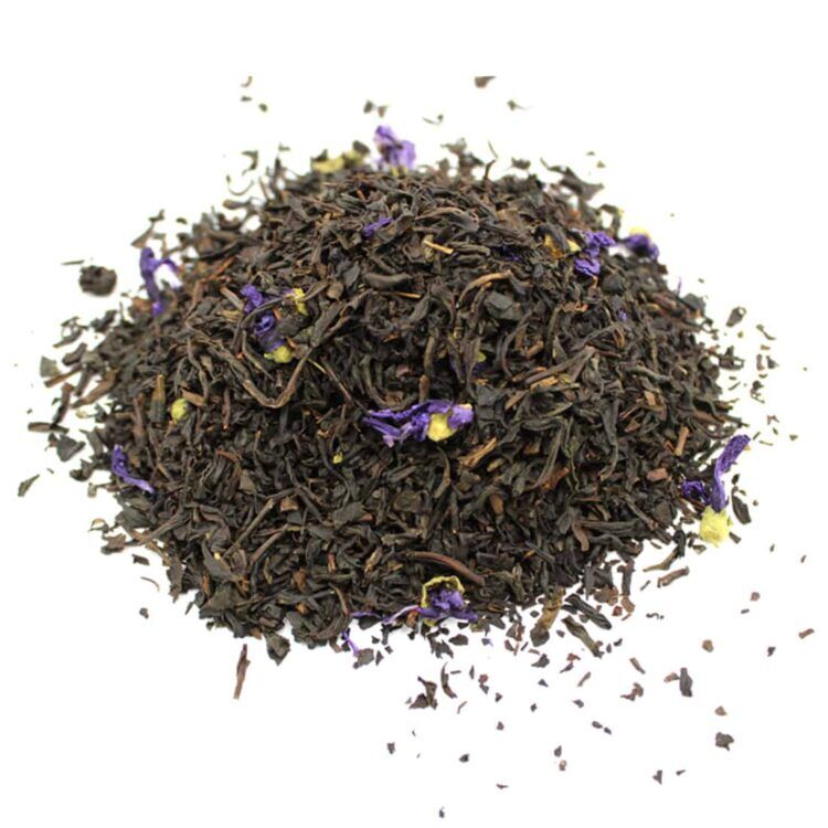 En klar plastbeholder indeholder løs te, som er en blanding af små, tørrede teblade. Tebladene er mørkebrune med nuancer af grå og grøn, og der er små, lyse blomster og citrusfrugtskaller blandet i. Etiket på beholderen er i en elegant skrifttype med sorte og guld nuancer, der angiver sorten "Merlin's Favorite Earl Grey".