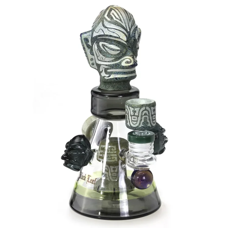 En diffusorbong i sort glas med en oval form pryder overfladen. Den har en glat, blank tekstur og er dekoreret med blade i et stiliseret mønster, der skaber en kontrast til det matte glas. Bongen har en krum hals og en bred bund, hvilket giver stabilitet. Det elegante design fremhæver formerne og detaljer i strukturen.