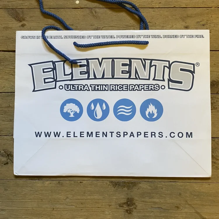 Papirhåndtasken har en rektangulær form med en glat overflade i en neutral farve. Tasken er prydet med subtile teksturer og en diskret, trykt logo på fronten. Håndtagene er lavet af kraftigt papir og er fastgjort med små, synlige metalclips. Taskens kanter er glatte og velformede, hvilket giver et elegant udtryk.