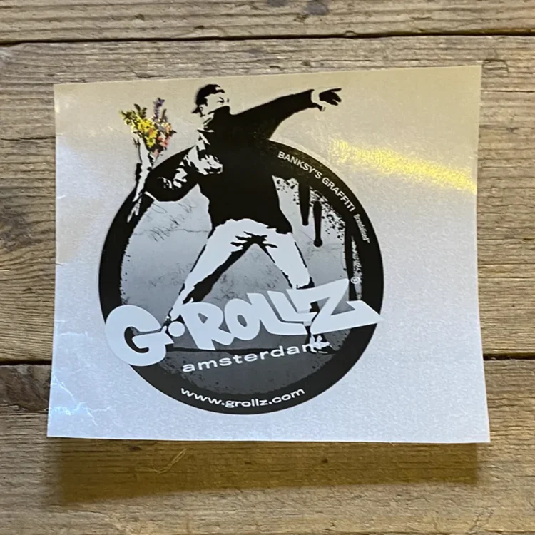 Klistermærket præsenterer et grafisk motiv med en rød blomst, der vokser ud af en grå mur. Blomsten er detaljeret med en grøn stilk og blade, hvor farverne står i kontrast til den matte baggrund. Mønsteret har en urban stil, der minder om street art, med tydelige konturer og en simpel, men stærk visuel appel.