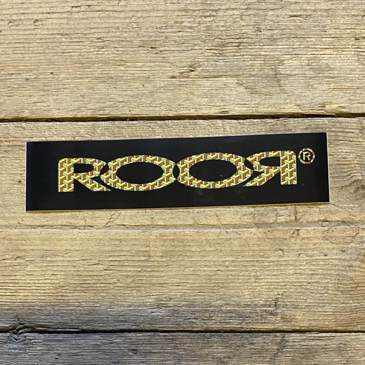 Rør-logoet fremtræder i en kontrastfuld kombination af sort og gul. Det sortfarvede område udgør baggrunden, mens det gule logo er designet med skarpe kanter og en tydelig skrifttype. Bogstaverne er store og klare, hvilket giver en markant visuel effekt. Materialet ser glat ud, hvilket indikerer en plastisk tekstur, ideel til klistermærker.