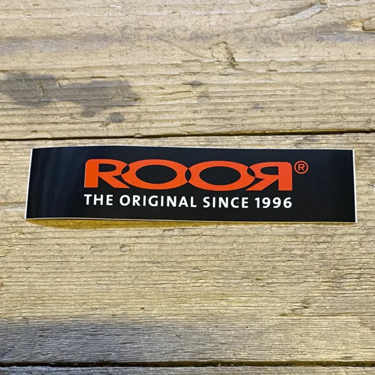 Et klistermærke i rektangulær form præsenterer et logo med en kombination af sort og rød farve. Logoet indeholder grafiske elementer og tekst i en moderne skrifttype. Den sorte baggrund fremhæver det røde design, hvilket skaber en kontrast. Klistermærket har en glat overflade med en let refleksion, der indikerer en plastisk kvalitet.