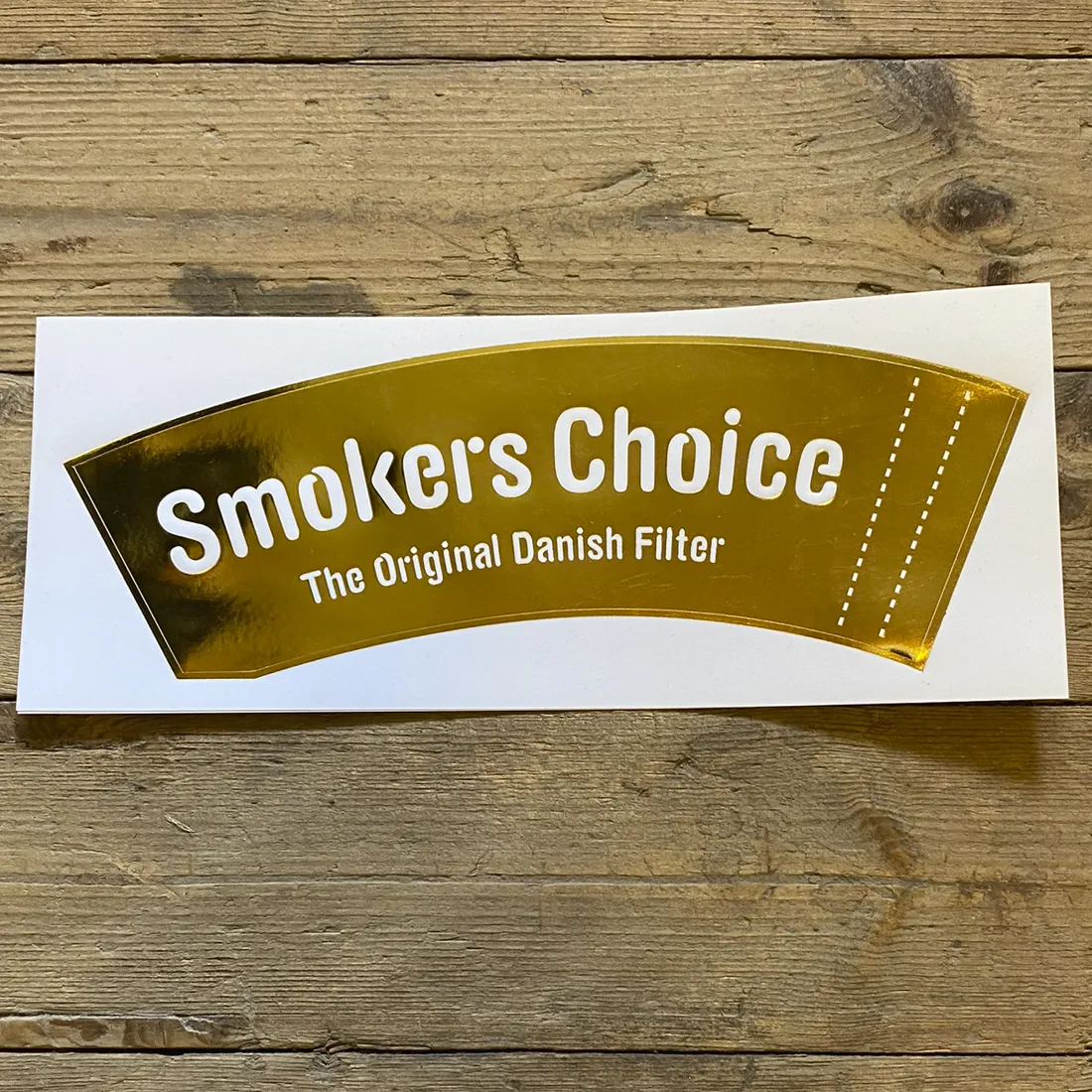 Klistermærket præsenterer et logo med teksten "Smoker's Choice" i store, elegante bogstaver. Farven er en dyb guld nuanceret tone, hvilket giver en skinnende og luksuriøs effekt. Baggrunden er ensfarvet, hvilket fremhæver logoets detaljer. Den glatte overflade af klistermærket skaber en refleksion, der tilføjer dybde til designet.