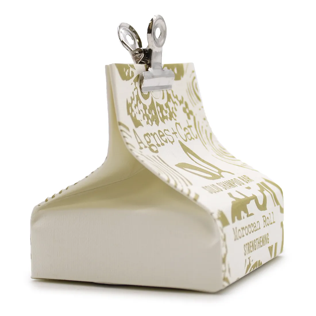 En solid shampoo bar præsenteres i en rund form med glatte kanter. Overfladen har en let tekstur, der fremstår mat og naturlig i en lys beige nuance. Den er forsynet med en lille, sort etiket, der angiver produktnavnet i hvid skrift. Shampoo baren hviler på en lys, træagtig overflade, som skaber en varm kontrast til produktet.