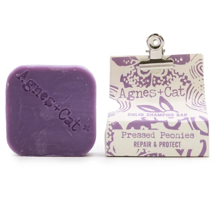 En solid shampoo-bar fremstår i en rund form, domineret af en blød beige farve med subtile nuancer. Overfladen er let struktureret, hvilket giver en mat finish. Der er indgraverede detaljer, som minder om blomstermønstre, der tilføjer en dekorativ dimension. Baren ligger på en naturlig, lys træoverflade, der kontrasterer med produktets farvetoner.