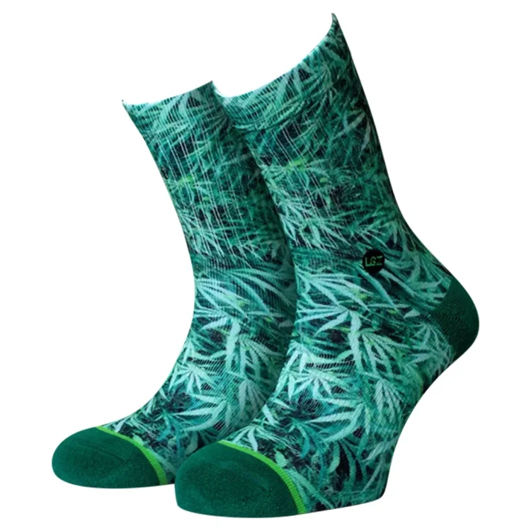 Sokkerne har en klassisk design med et mønster af bladformer i nuancer af grøn, gul og brun. Baggrunden er i en lys grå farve, der fremhæver mønsteret. Materialet ser blødt og strækbart ud, med en let ribbet struktur ved toppen. Kanten er ensfarvet og afslutter sokken med en glat finish.