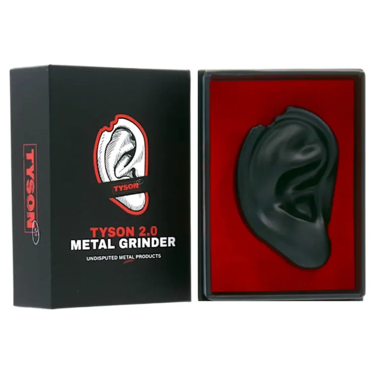 En sort metal grinder præsenterer sig med en glat, mat overflade. Den runde form har en let skrånende kant og er opdelt i to sektioner med en central, tandet mekanisme til formaling. Øverst er der en lille åbning, der muliggør påfyldning, mens en fin gitterstruktur i bunden sikrer filtrering. Kanten har en diskret, præcis afrunding.