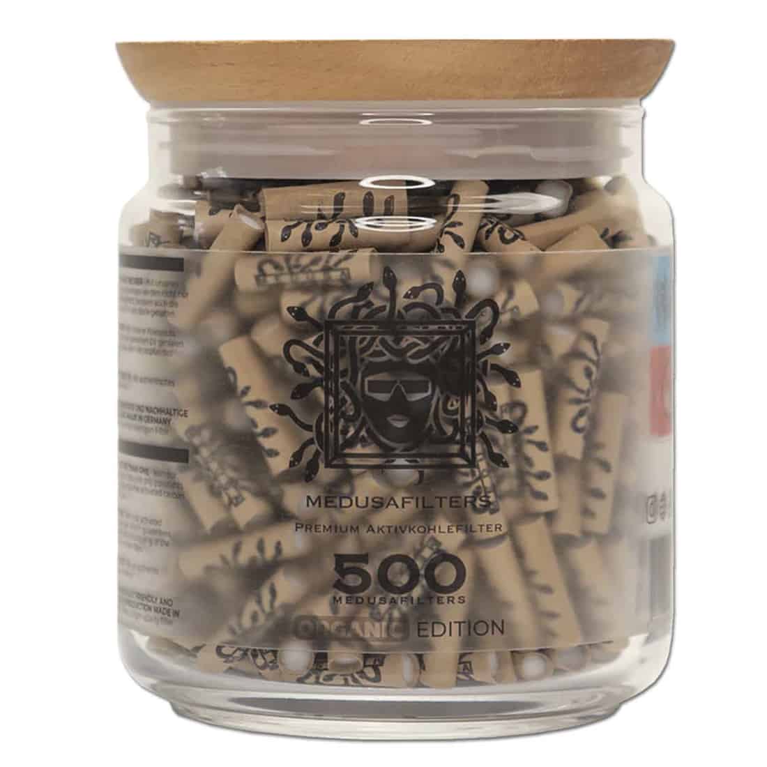 En pakke med 500 aktive kulfiltertips præsenteres i en rektangulær, gennemsigtig plastbeholder. Tipsene er sorte med en glat overflade, og de har en let skinnende tekstur. Beholderen har en etikette med grøn tekst og symboler, der angiver økologisk indhold. Låget er skruefunktionelt og sikrer tæt lukning. Mønstret på etiketten er enkelt og moderne.