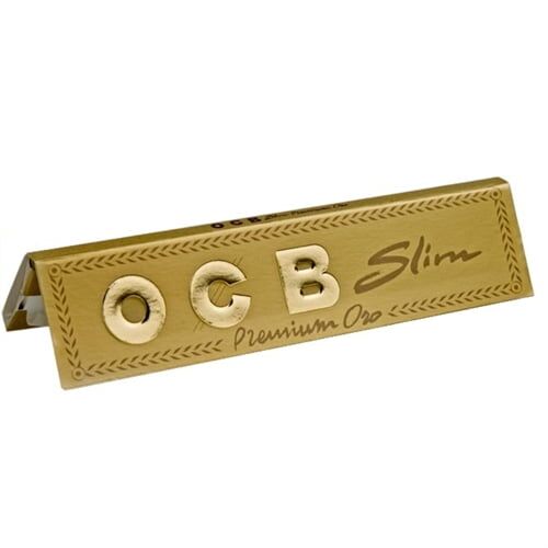 OCB Kingsize Slim Gold er en pakke med en gylden overflade, dekoreret med sort tekst og et stiliseret logo. Pakken har en rektangulær form med en åbningsklap i toppen. Den gyldne farve skaber en glitrende effekt, mens den sorte skrift kontrasterer tydeligt. Pakken indeholder slanke papirruller, der er synlige ved en let åbning i kanten.