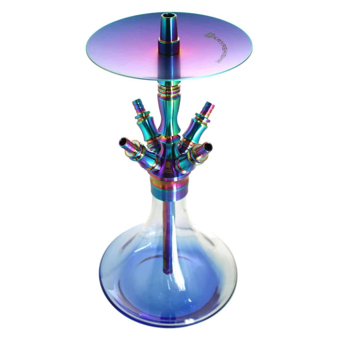 En rund skål med en glat, reflekterende overflade dominerer kompositionen. Skålen har en dyb blå farve med nuancer af lilla, der skaber en regnbueeffekt. Den er lavet af rustfrit stål og har en let bøjning ved kanten, hvilket giver et stilfuldt udtryk. Indvendigt er skålen glansfuld, mens ydersiden har en mere mat finish.