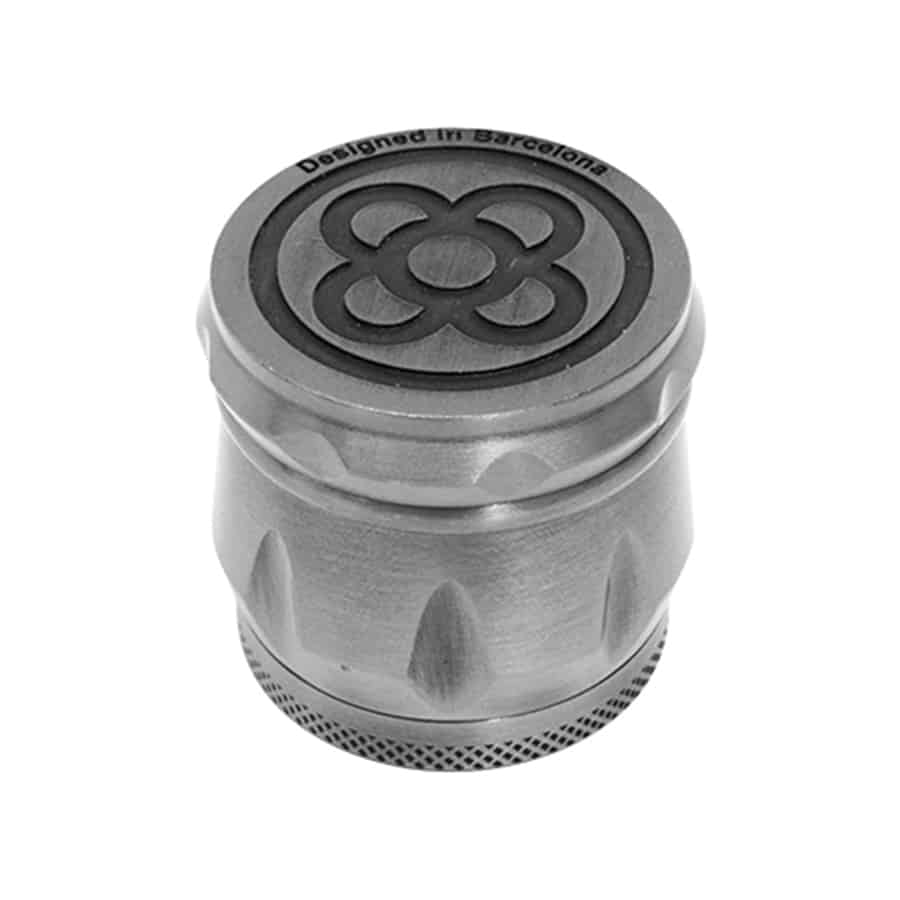 En sølvfarvet kværn med en blomsterpræget overflade. Kværnen har en rund form med en glat, metallisk finish, der reflekterer lys. De udskårne blomsterformer er detaljerede og giver en taktil tekstur. Kværnen består af flere lag, adskilt af fine, tætsiddende tænder, og har en skruelås øverst for lukning.