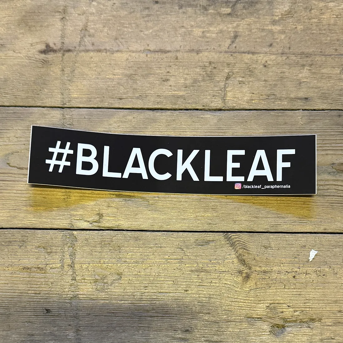 Et sort klistermærke prydes af et stiliseret bladdesign med detaljerede nervestrukturer. Overfladen er glat og skinnende, hvilket giver en let refleksion. Bladets konturer er skarpe, og dets form er asymmetrisk, hvilket skaber en dynamisk visuel effekt. Baggrunden er ensfarvet, hvilket fremhæver klistermærkets design og farve.