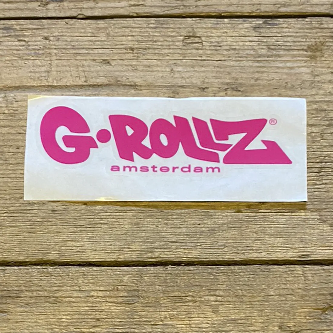 Klistermærket præsenterer et cirkulært design med en lys pink baggrund. I midten er der et stiliseret logo, der indeholder tekst og grafiske elementer, fremhævet af en kontrasterende farve. Teksten er sort, med en moderne skrifttype, der giver et klart og tydeligt udtryk. Overfladen har en glat, skinnende finish, der reflekterer lys.