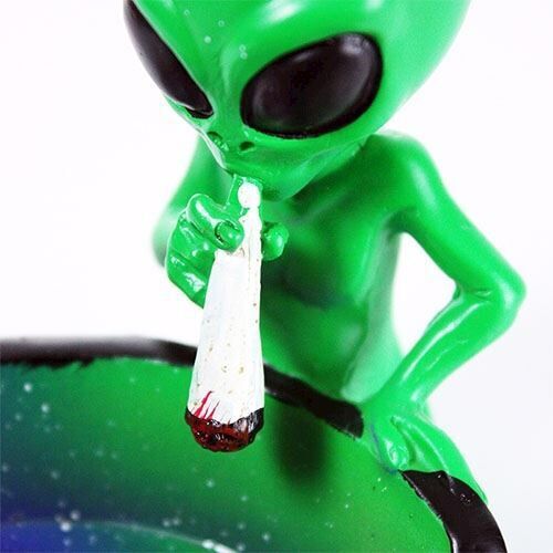 Et askebæger i høj, oval form er fremstillet af transparent, grønligt glas. Overfladen har en glat tekstur med en let bølget kant. Bunden er flad og stabil, mens der er en lille indgravering, der fungerer som et dekorativt element. Farvevariationer ses i lyset, der reflekteres gennem glasset, hvilket skaber en subtil, æstetisk effekt.