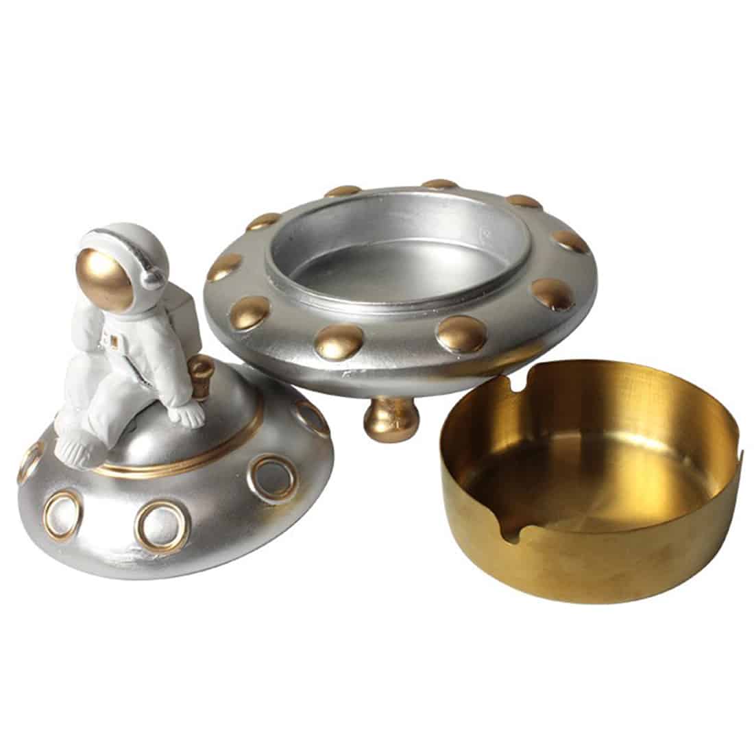 En ashytronaut med detaljeret udformning, der ligner en astronaut i rumdragt. Figuren har en blank, hvid overflade med sorte detaljer, såsom visir og søm. Under figuren er en lille rumskibslignende beholder, der fungerer som askebæger, med en grå, metallisk finish. Den skulpturelle form skaber en interessant kontrast mellem det bløde og hårde materiale.