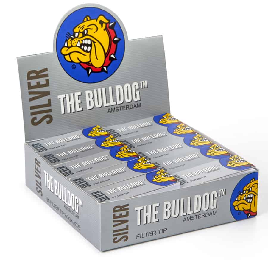 En rektangulær boks i metal med en glinsende sølvfarve, prydet med sort tekst og grafik. Boksen har afrundede hjørner og en let mat finish på bagsiden. Fronten indeholder et logo og informationstekst i hvid og sort. Låget er let buet, og der er en synlig åbning i den ene ende, der antyder funktionalitet.