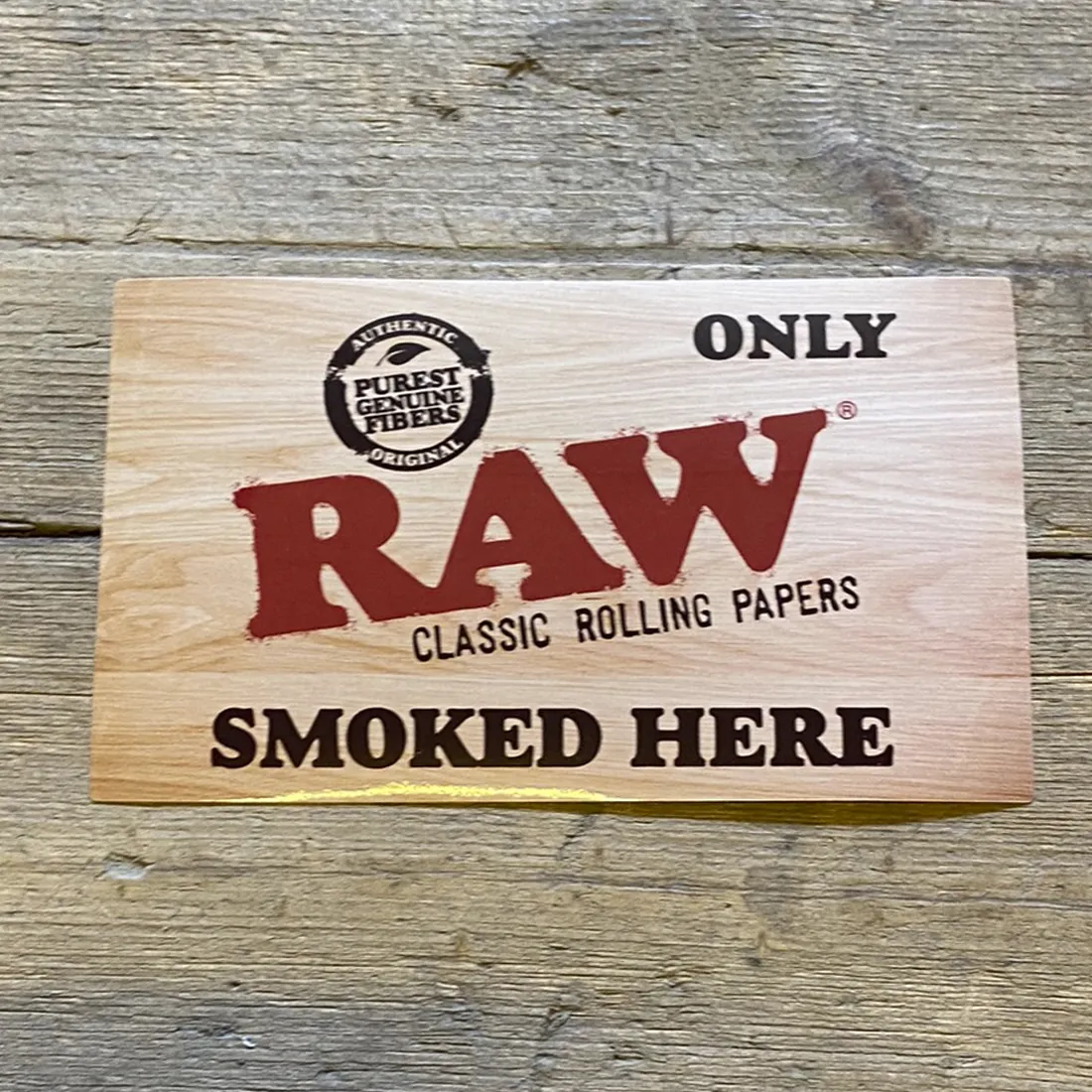 Klistermærket har en rektangulær form med en mat, sort baggrund. Øverst fremtræder teksten "RAW SMOKED" i store, hvide, sans-serif bogstaver, mens "HERE" er placeret under i mindre størrelse. Teksten har en let slidt effekt, hvilket giver et rustikt udtryk. Der er ingen yderligere grafiske elementer eller farver på klistermærket.