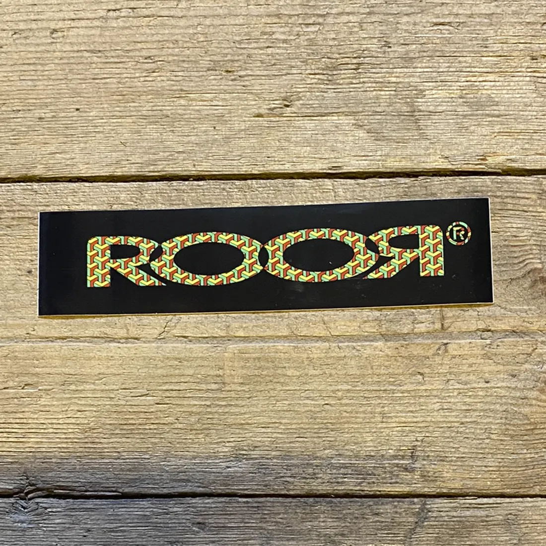 Rør-logoet fremtræder i en kontrastfuld kombination af sort og gul. Det sortfarvede område udgør baggrunden, mens det gule logo er designet med skarpe kanter og en tydelig skrifttype. Bogstaverne er store og klare, hvilket giver en markant visuel effekt. Materialet ser glat ud, hvilket indikerer en plastisk tekstur, ideel til klistermærker.